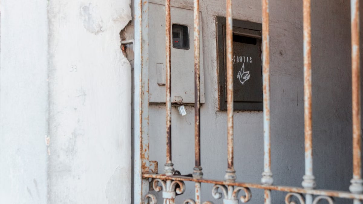 A rusty metal fence frames an old mail drop against a worn wall, creating a rustic atmosphere.