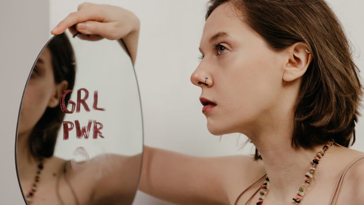 A young woman gazes into a mirror with 'GRL PWR' written on it, emphasizing empowerment.
